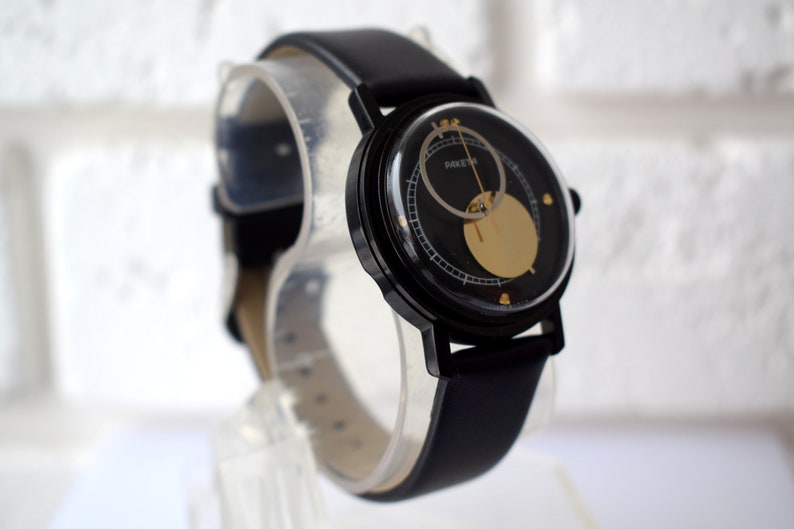 May include: A black wristwatch with a black leather band and a circular face. The face has a gold-colored circle in the center and a smaller circle around it. The watch face also has black markings and the word "RAKET" printed on it.
