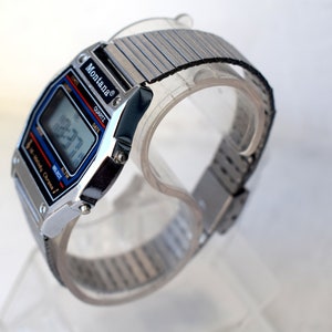 K&ouml;nnte beinhalten: Silberfarbene Montana Digitaluhr mit schwarzem und blauem Zifferblatt, das die Uhrzeit anzeigt. Die Uhr hat ein dehnbares silbernes Armband. Die Uhr wird auf einem transparenten St&auml;nder pr&auml;sentiert.
