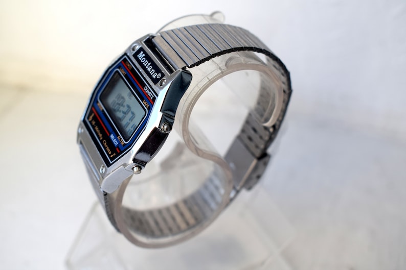 K&ouml;nnte beinhalten: Silberfarbene Montana Digitaluhr mit schwarzem und blauem Zifferblatt, das die Uhrzeit anzeigt. Die Uhr hat ein dehnbares silbernes Armband. Die Uhr wird auf einem transparenten St&auml;nder pr&auml;sentiert.