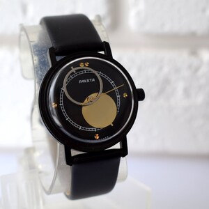 May include: A black wristwatch with a black leather band. The watch face is black with a gold circle and a silver ring around the edge. The watch face also has the word "PAKEta" printed on it.