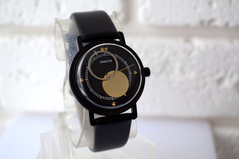 May include: A black wristwatch with a black leather band. The watch face is black with a gold circle and a silver ring around the edge. The watch face also has the word "PAKEta" printed on it.