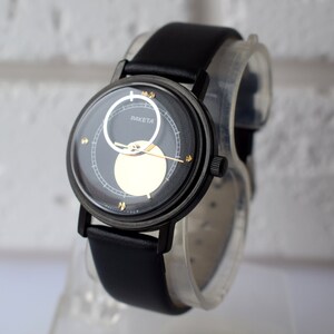 May include: A black wristwatch with a black leather band. The watch face has a black background with gold accents and a white circle in the center. The watch face also has the word "RAKETTA" printed on it.