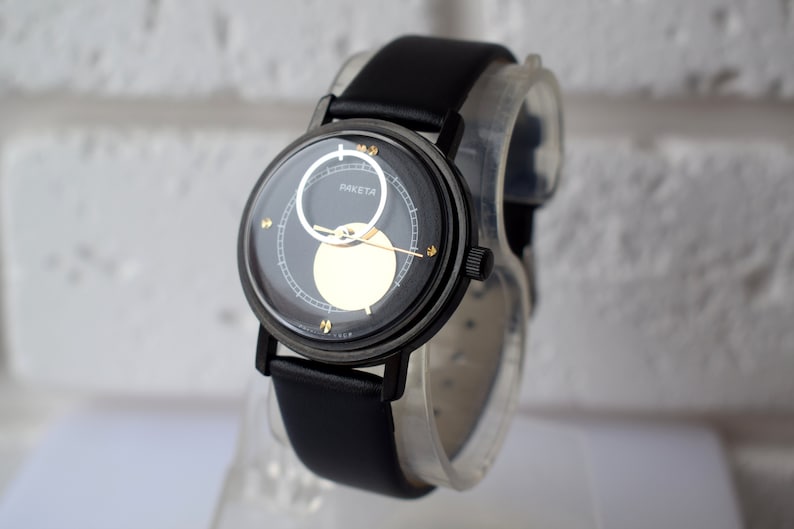 May include: A black wristwatch with a black leather band. The watch face has a black background with gold accents and a white circle in the center. The watch face also has the word "RAKETTA" printed on it.