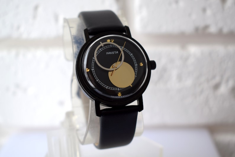 May include: A black wristwatch with a black leather band and a round face. The face has a gold circle in the center and the word "RAKET" printed on it.