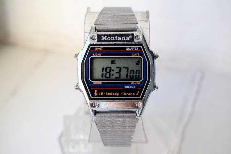 K&ouml;nnte beinhalten: Silberne Montana Digitaluhr mit rechteckigem Zifferblatt. Die Uhr zeigt Zeit, Datum und andere Funktionen auf einem digitalen Bildschirm an. Sie hat ein silbernes Metallarmband und den Text "Montana" und "16-Melody Chrono".