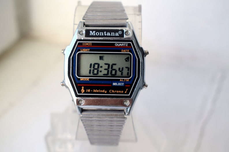 K&ouml;nnte beinhalten: Silberfarbene Montana Quartz Digitaluhr mit rechteckigem Zifferblatt. Die Uhr zeigt Uhrzeit, Datum und andere Funktionen auf einem digitalen Bildschirm an. Das Armband besteht aus silberfarbenen Metallgliedern. Auf dem Zifferblatt steht "Montana", "Quartz" und "16-Melody Chrono".