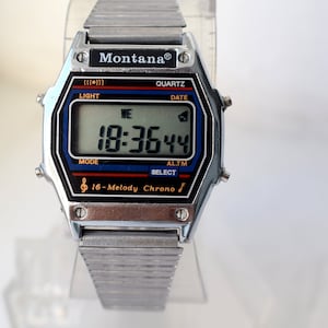 K&ouml;nnte beinhalten: Eine silberfarbene Montana Digitaluhr mit rechteckigem Zifferblatt, das die Uhrzeit 18:36:44 anzeigt. Das Zifferblatt hat schwarze, blaue und rote Akzente mit dem Text "16-Melody Chrono". Das Armband besteht aus silberfarbenem Metall.