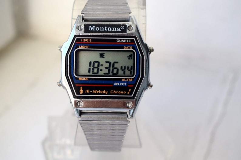 K&ouml;nnte beinhalten: Eine silberfarbene Montana Digitaluhr mit rechteckigem Zifferblatt, das die Uhrzeit 18:36:44 anzeigt. Das Zifferblatt hat schwarze, blaue und rote Akzente mit dem Text "16-Melody Chrono". Das Armband besteht aus silberfarbenem Metall.