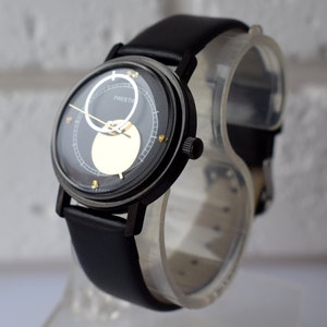 May include: A black wristwatch with a black leather band. The watch face has a black background with a gold circle and a white circle with the word "RAKET" printed on it.