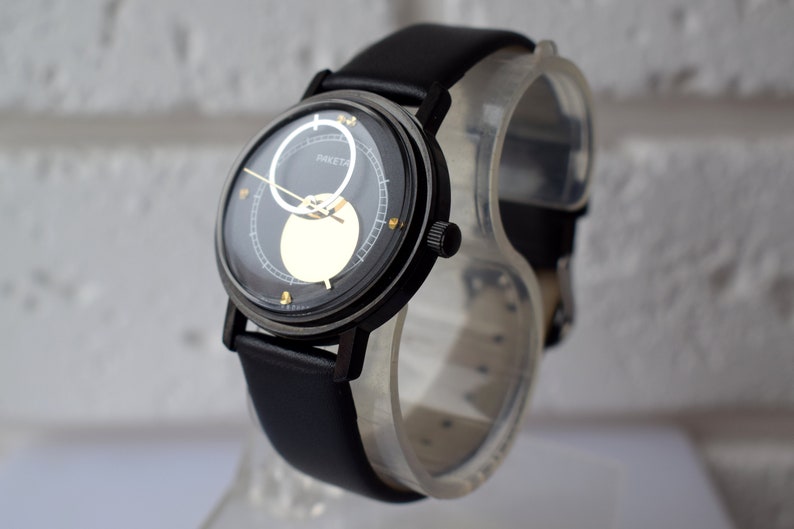 May include: A black wristwatch with a black leather band. The watch face has a black background with a gold circle and a white circle with the word "RAKET" printed on it.