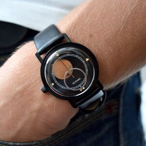May include: A black wristwatch with a black leather band and a circular face. The face has a black background with a gold ring and a gold circle in the center. The word "PARKER" is printed in white on the face.