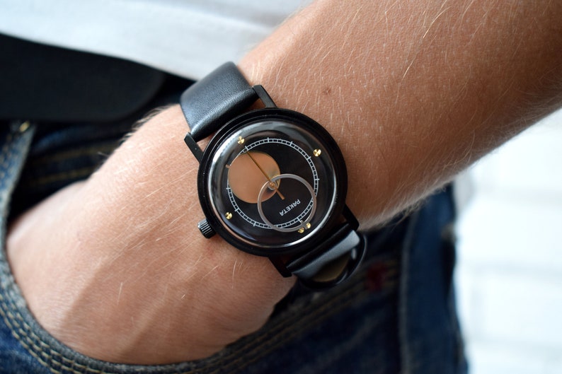May include: A black wristwatch with a black leather band and a circular face. The face has a black background with a gold ring and a gold circle in the center. The word "PARKER" is printed in white on the face.