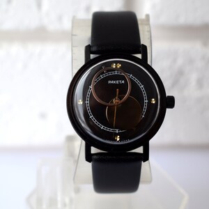 May include: A black wristwatch with a black leather band and a circular face. The face has a gold ring around the edge and a smaller circle in the center with the word "PAKETA" printed on it.