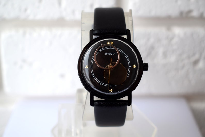 May include: A black wristwatch with a black leather band and a circular face. The face has a gold ring around the edge and a smaller circle in the center with the word "PAKETA" printed on it.