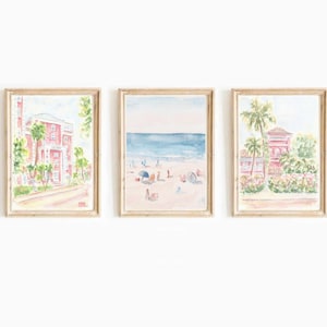 May include: Set of three framed watercolor prints. The prints depict beach scenes and buildings with a soft, pastel color palette. The artwork includes a beach scene with people, a building with palm trees, and a building on a street.