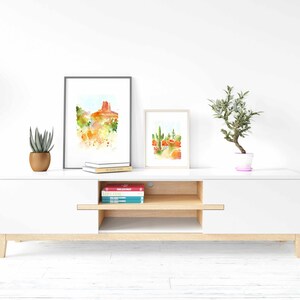 May include: A white entertainment center with a wood top and a single drawer. The entertainment center has two shelves and is decorated with two framed watercolor paintings of desert landscapes and a potted plant.