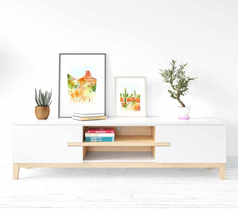 May include: A white entertainment center with a wood top and a single drawer. The entertainment center has two shelves and is decorated with two framed watercolor paintings of desert landscapes and a potted plant.