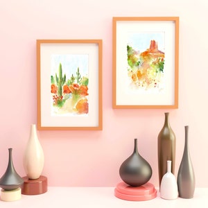 May include: Two framed watercolor paintings of desert landscapes, one with a cactus and the other with a red rock formation.  The paintings are hanging on a light pink wall with six decorative vases in front of them.