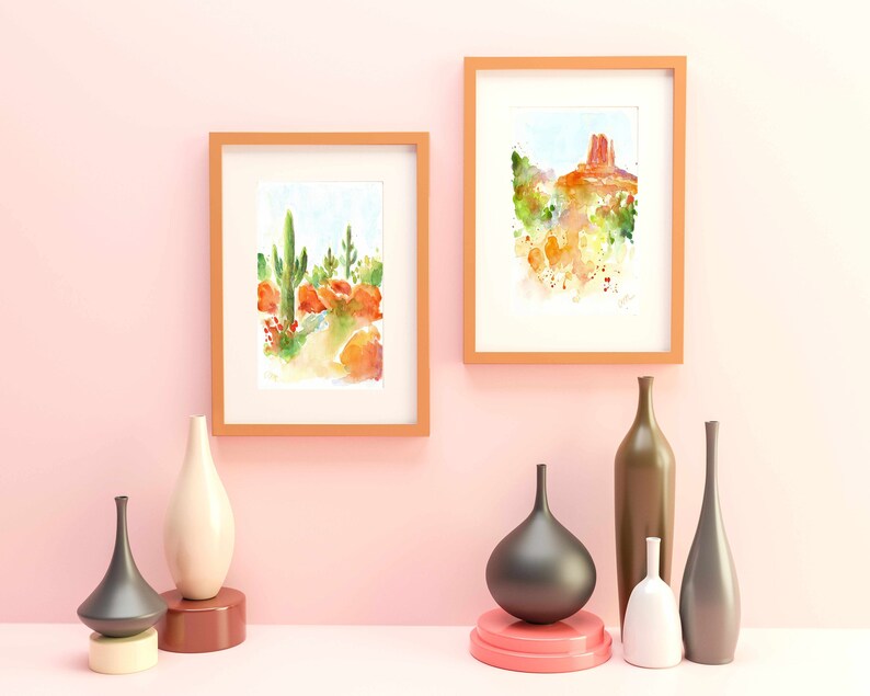 May include: Two framed watercolor paintings of desert landscapes, one with a cactus and the other with a red rock formation.  The paintings are hanging on a light pink wall with six decorative vases in front of them.