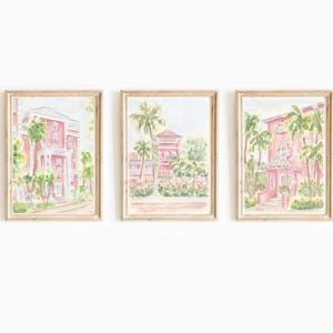 May include: Set of three framed watercolor paintings featuring pink buildings with palm trees. The artwork has a light, airy feel, with soft colors and a focus on architectural details. The frames are a light wood color.