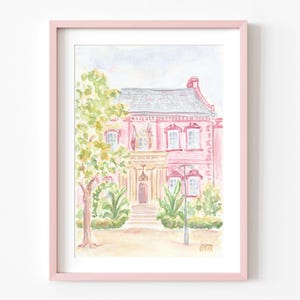 May include: Watercolor painting of a pink house with a white door and windows. The house is surrounded by green trees and bushes. The painting is framed in a light pink frame.