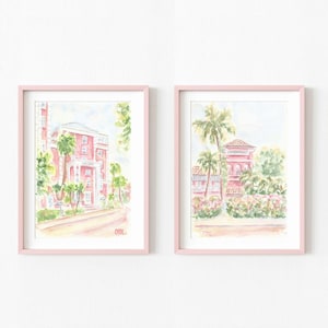 May include: Two framed watercolor prints featuring pink buildings with palm trees and lush greenery. The artwork is set against a white background, with each print encased in a light pink frame. The style is reminiscent of a tropical resort.
