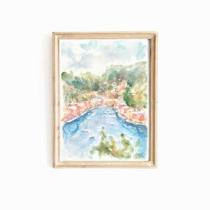 May include: A watercolor painting of a coastal town with colorful buildings and a blue bay. The painting is framed in a light wood frame.