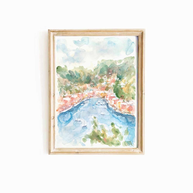 May include: A watercolor painting of a coastal town with colorful buildings and a blue bay. The painting is framed in a light wood frame.