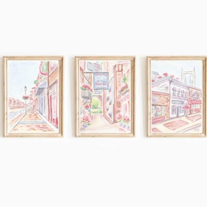 May include: Set of three framed watercolor paintings depicting quaint street scenes. The artwork features buildings with awnings, flower boxes, and a sign that reads "Blues". The color palette includes soft pinks, oranges, and blues, creating a charming, vintage aesthetic.
