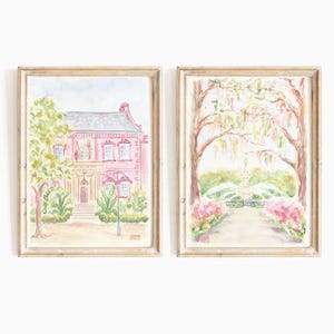 May include: Two watercolor paintings in light wood frames. The left painting shows a pink house with white trim and green trees. The right painting shows a path leading to a fountain in a park with pink flowers and trees.