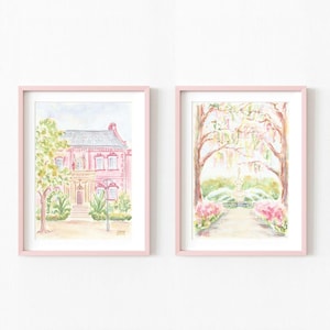 Op de afbeelding: Twee aquarellen in roze lijsten. De eerste afbeelding toont een roze huis met een gele deur en groene bomen. De tweede afbeelding toont een pad dat naar een fontein met bomen en bloemen leidt.