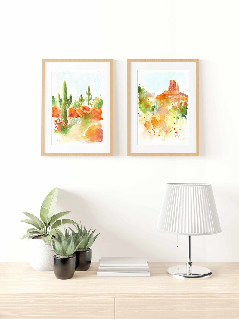 May include: Two framed watercolor paintings of desert landscapes. The first painting shows a tall cactus in the foreground with red rock formations in the background. The second painting shows a red rock formation with a blue sky in the background.