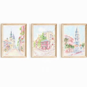 May include: A set of three watercolor paintings depicting scenes from a city. The first painting shows a street with a church in the background. The second painting shows a street with a pink house in the background. The third painting shows a street with a tall steeple in the background.