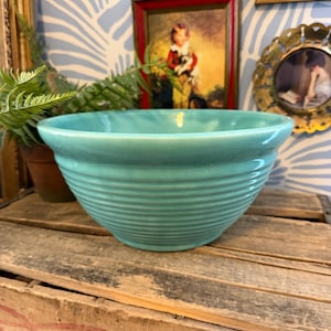 May include: A turquoise ceramic bowl with horizontal ribbed detailing. The bowl has a wide, shallow shape and a smooth, glossy finish. It sits on a wooden crate.
