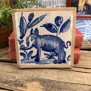 May include: Square ceramic tile with a hand-painted blue elephant design. The elephant is surrounded by blue foliage against a white background. The tile is set in a wooden frame, creating a decorative piece.
