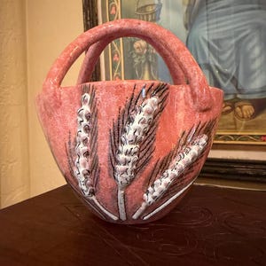 May include: A coral-colored ceramic basket with a handle, featuring a wheat stalk design in white and black. The basket has a textured surface and is likely vintage or antique.