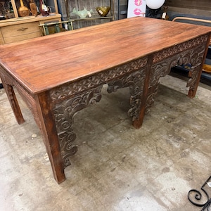 May include: A rectangular wooden table with a smooth, medium-brown top and intricate, dark-stained carved detailing along the apron and legs. The table features ornate, scroll-like carvings that extend from the frame down to the four sturdy legs.