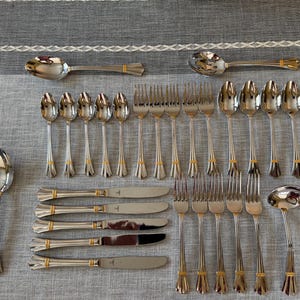 May include: A stainless steel flatware set featuring silver-toned utensils with gold-colored accents at the base of the handles. The collection includes dinner knives, forks, spoons, a soup ladle, and a small shell-patterned spoon, all arranged in rows.