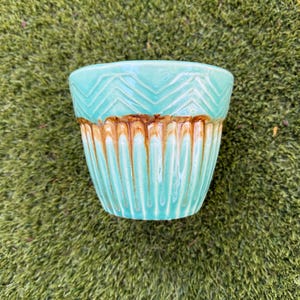 May include: A turquoise ceramic planter with a brown band and vertical grooves. The top section features a zigzag pattern. This decorative pot is suitable for small plants and adds a touch of color to any space.