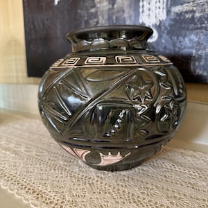 May include: A dark green ceramic vase with intricate embossed designs, including stars and geometric patterns. The vase has a rounded shape with a narrow neck and a decorative rim. The base features a pink and white design.