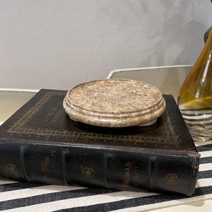 May include: A beige, circular decorative object with a textured surface sits atop a dark brown book. The book has gold lettering and is placed on a black and white striped cloth. A glass bottle is visible in the background.