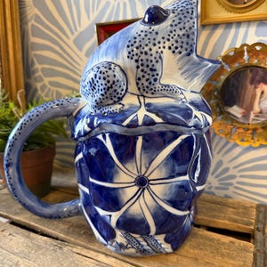 May include: A blue and white ceramic pitcher with a frog-shaped lid. The pitcher features a detailed floral pattern and a curved handle. The frog has dark blue spots and is perched on top of the pitcher. Decorative home decor.