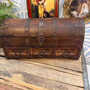 May include: A vintage wooden chest with three small drawers and metal ring pulls. The chest is dark brown with metal bands and a clasp. The chest sits on a wooden crate.