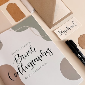 May include: A white and brown notebook with the title "An Introduction to Brush Calligraphy with Blissfully Yours" in black script. The notebook is surrounded by craft supplies, including brown paper tags, a white envelope, and black calligraphy pens.