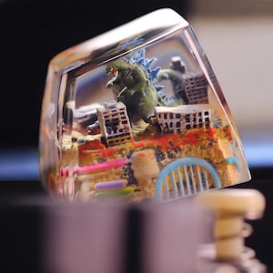 May include: A clear resin keycap featuring a miniature diorama of a city under attack by a green Godzilla-like monster. The scene includes detailed buildings, colorful accents, and a blue monster. The keycap is designed for mechanical keyboards.