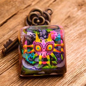 May include: A colorful, square resin keycap featuring a Majora's Mask design. The keycap is filled with a miniature landscape of flowers, rocks, and a mountain. A bronze-colored key and decorative element are in the background, set on a wooden surface.