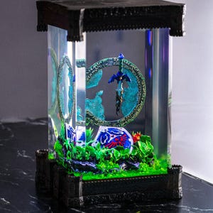 Op de afbeelding: Een decoratieve display case met een fantasiethema diorama. De case heeft een zwart frame en een heldere acryl behuizing. Binnenin toont een miniatuurlandschap met een blauw en groen kleurenschema een zwaard- en schildontwerp.