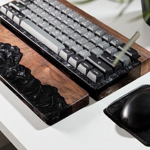 Puede incluir: Un teclado mecánico personalizado con teclas grises, sobre un reposamuñecas de madera con una incrustación de resina negra. También hay un reposamuñecas a juego y un ratón negro con una alfombrilla de cuero.