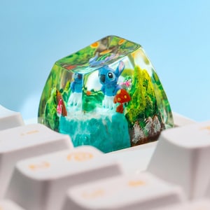 May include: A custom keycap featuring a miniature diorama with a blue alien character, a mushroom, and green foliage. The keycap is clear, allowing a view of the detailed scene inside. The keycap is set on a white keyboard.