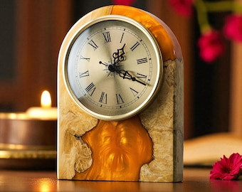 May include: A decorative wooden clock with a gold-colored face and Roman numerals. The clock has a unique design with a light-colored wood base and a gold-colored resin accent. The clock is a stylish home decor item.